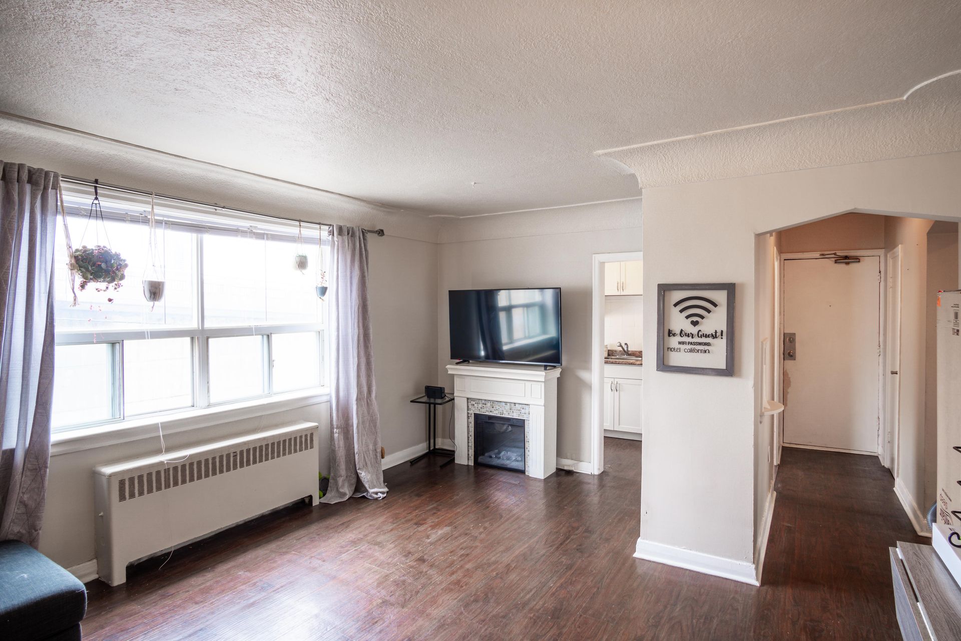 Bright living room with large window, radiator, TV on mantel, and hallway to the right