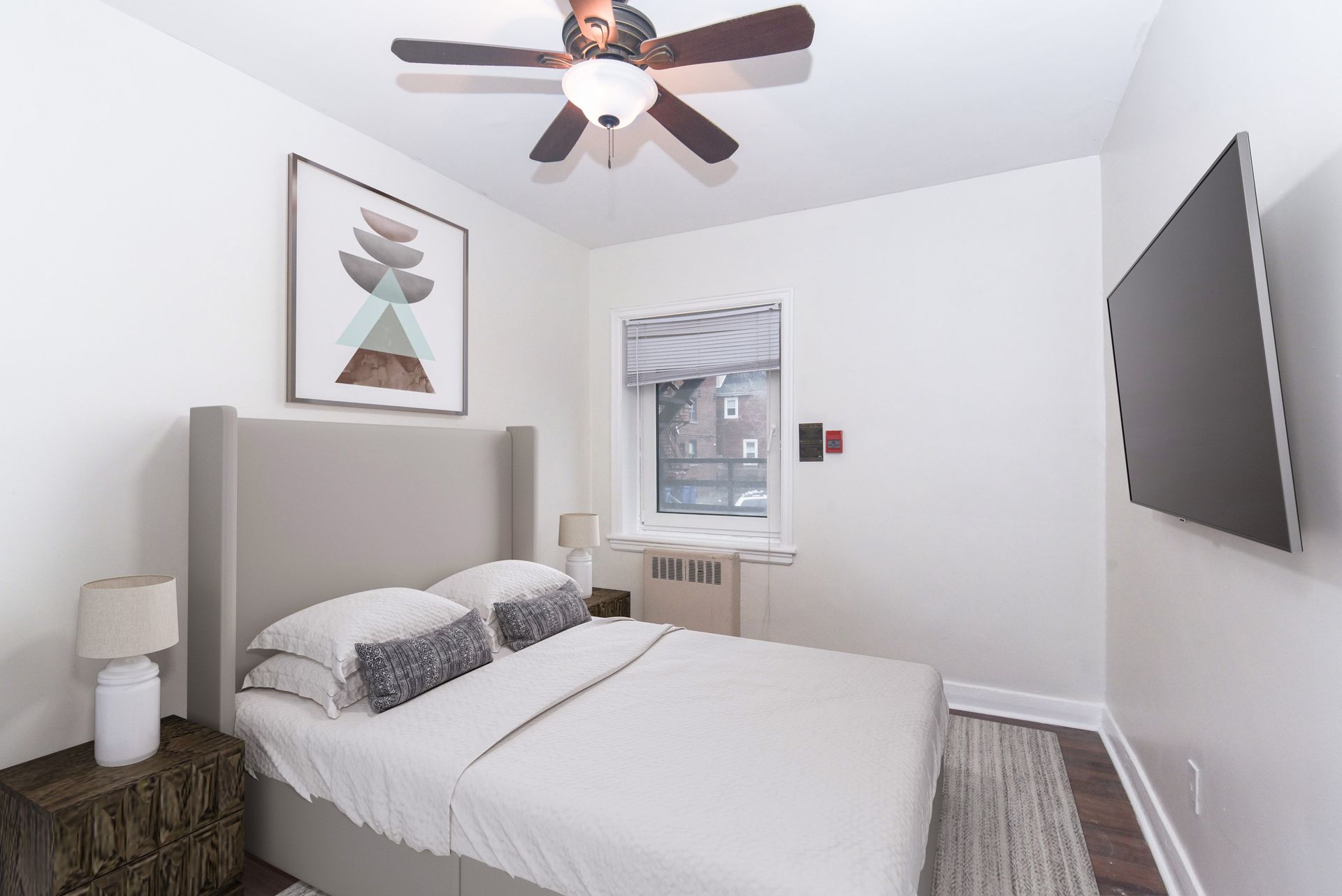 Bright bedroom with a bed, wall art, ceiling fan, window, and wall-mounted TV.