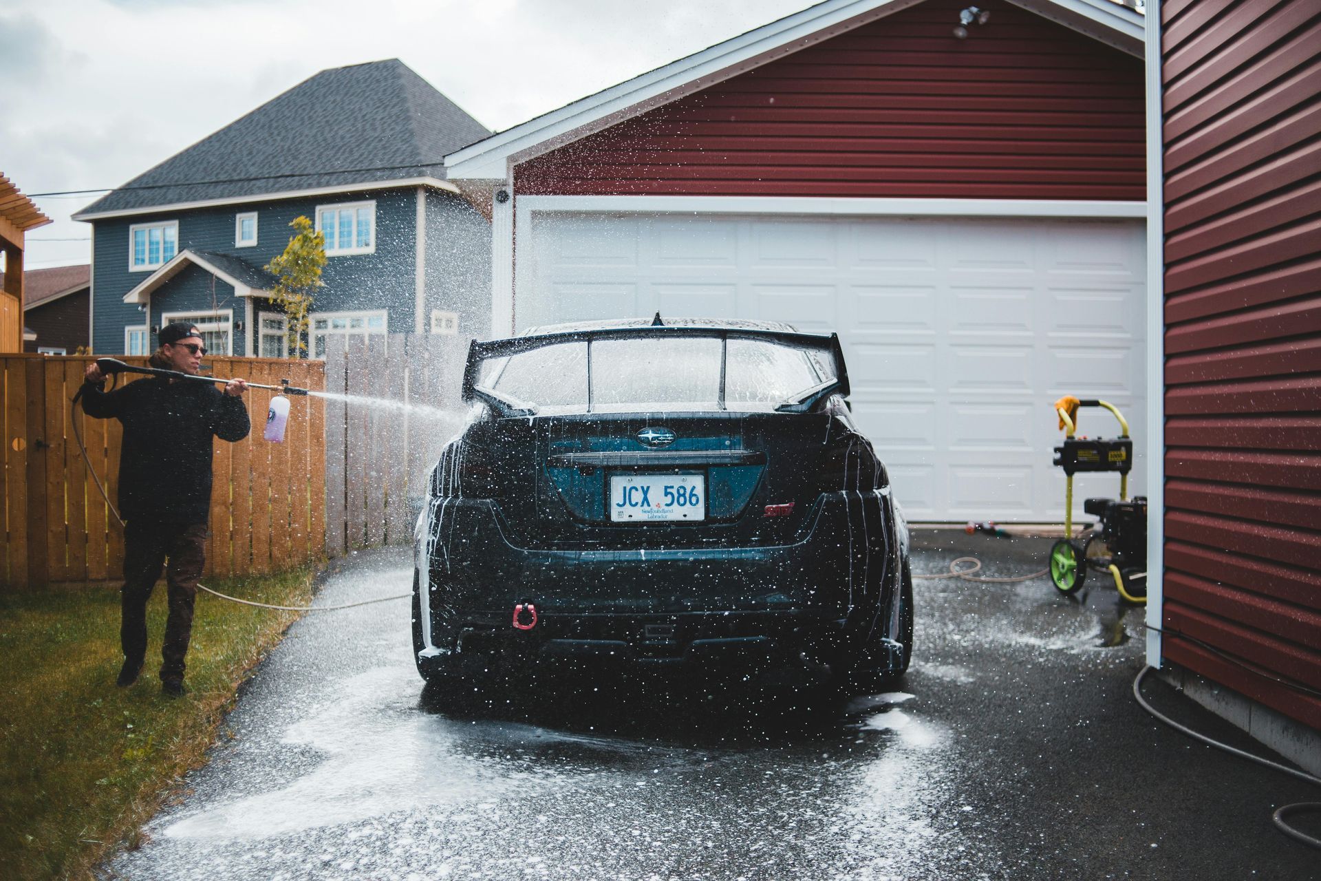 Person washing a black car in a driveway with a hose, beside a garage on a rainy day.