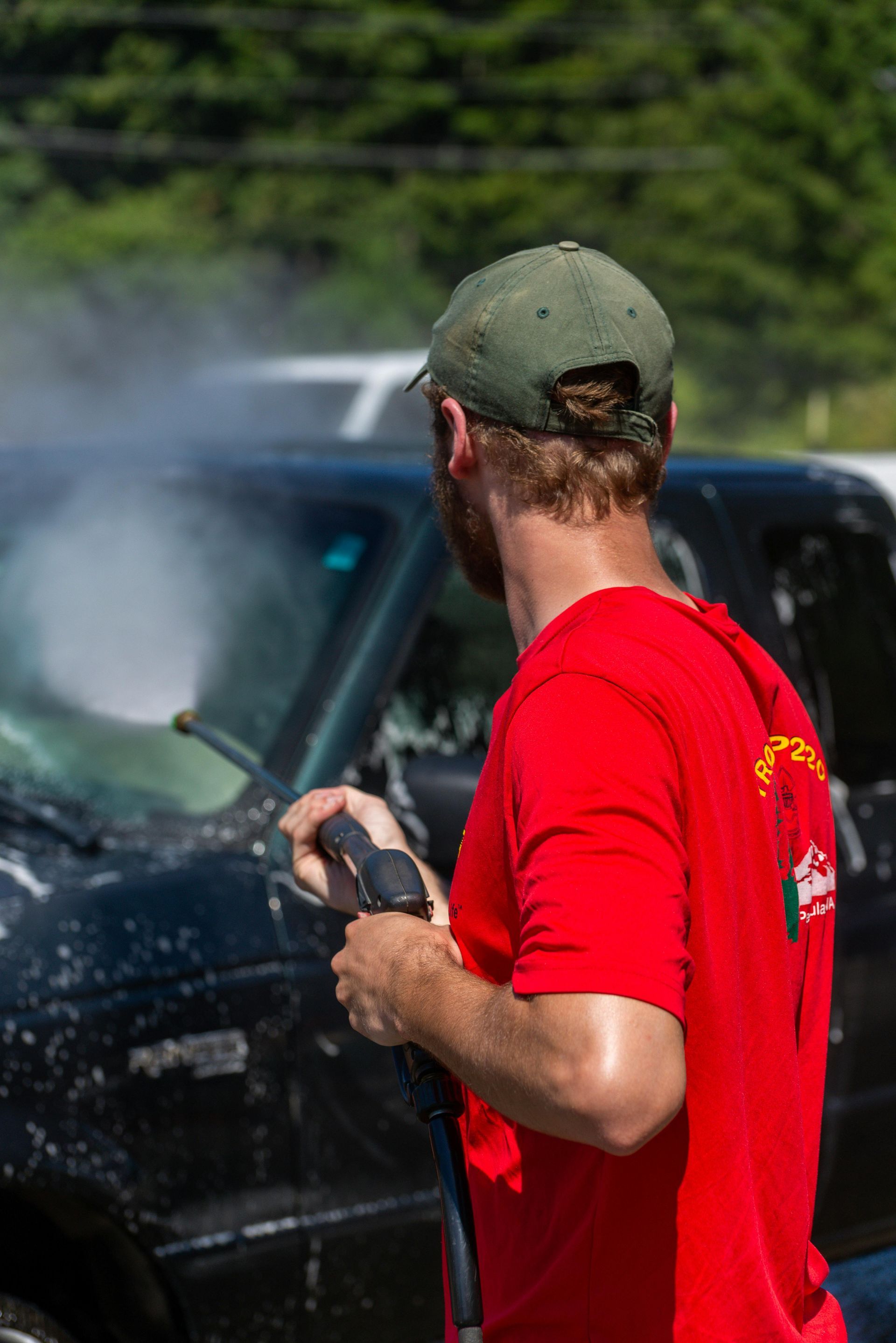 Person in red shirt spraying water on a black vehicle outdoors