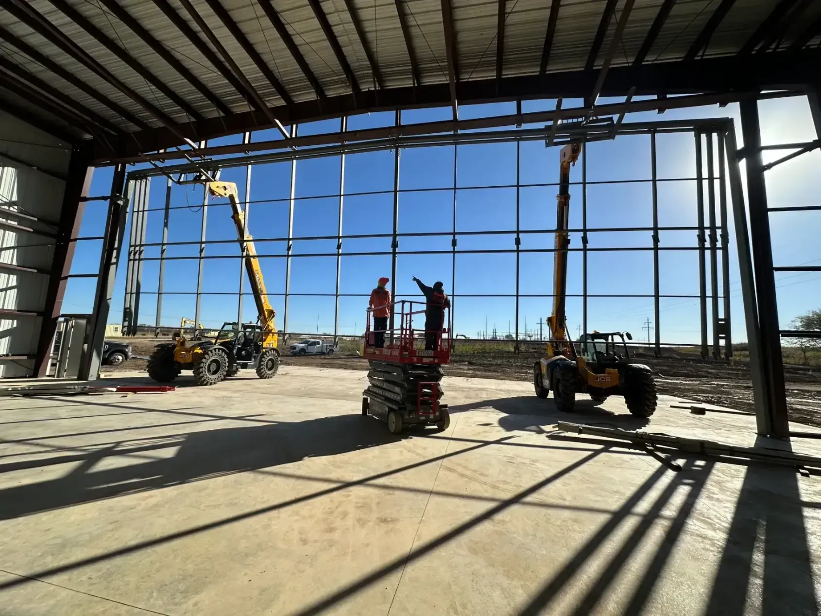 Construction workers on lifts inside a large building frame, installing window supports. Bright sun, blue sky.