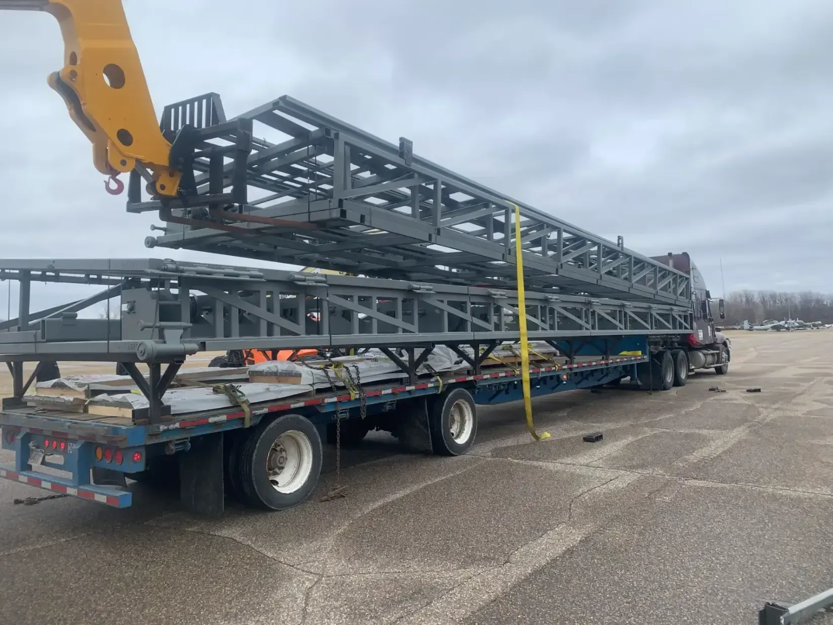 A flatbed truck carrying gray metal framework, being loaded by a yellow crane arm in a parking lot.