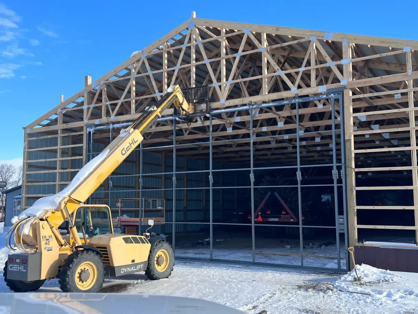 Yellow telehandler at construction site of a wooden framed building in snow, clear blue sky.