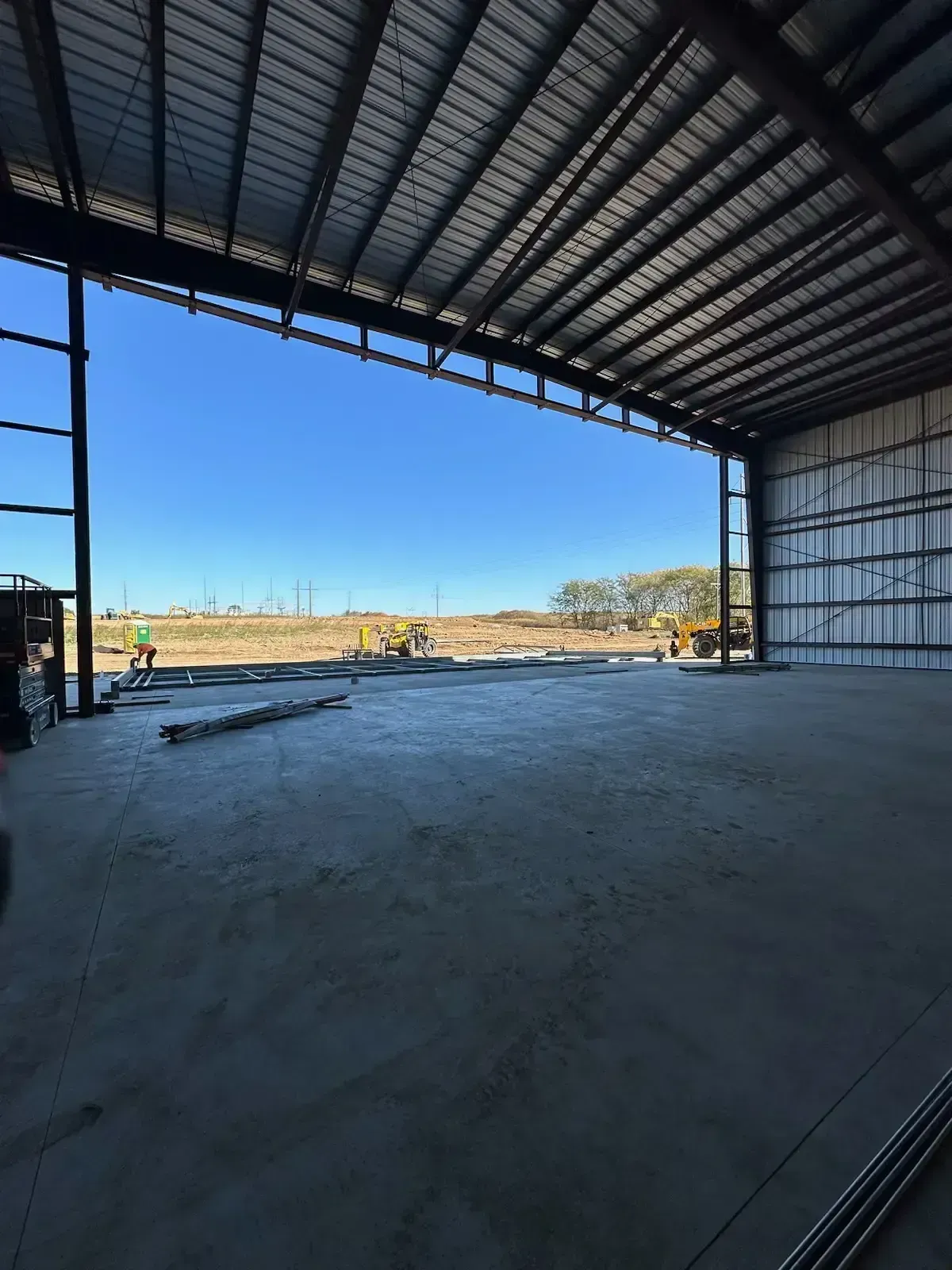 Inside a large metal building, concrete floor, open to a construction site, blue sky visible.
