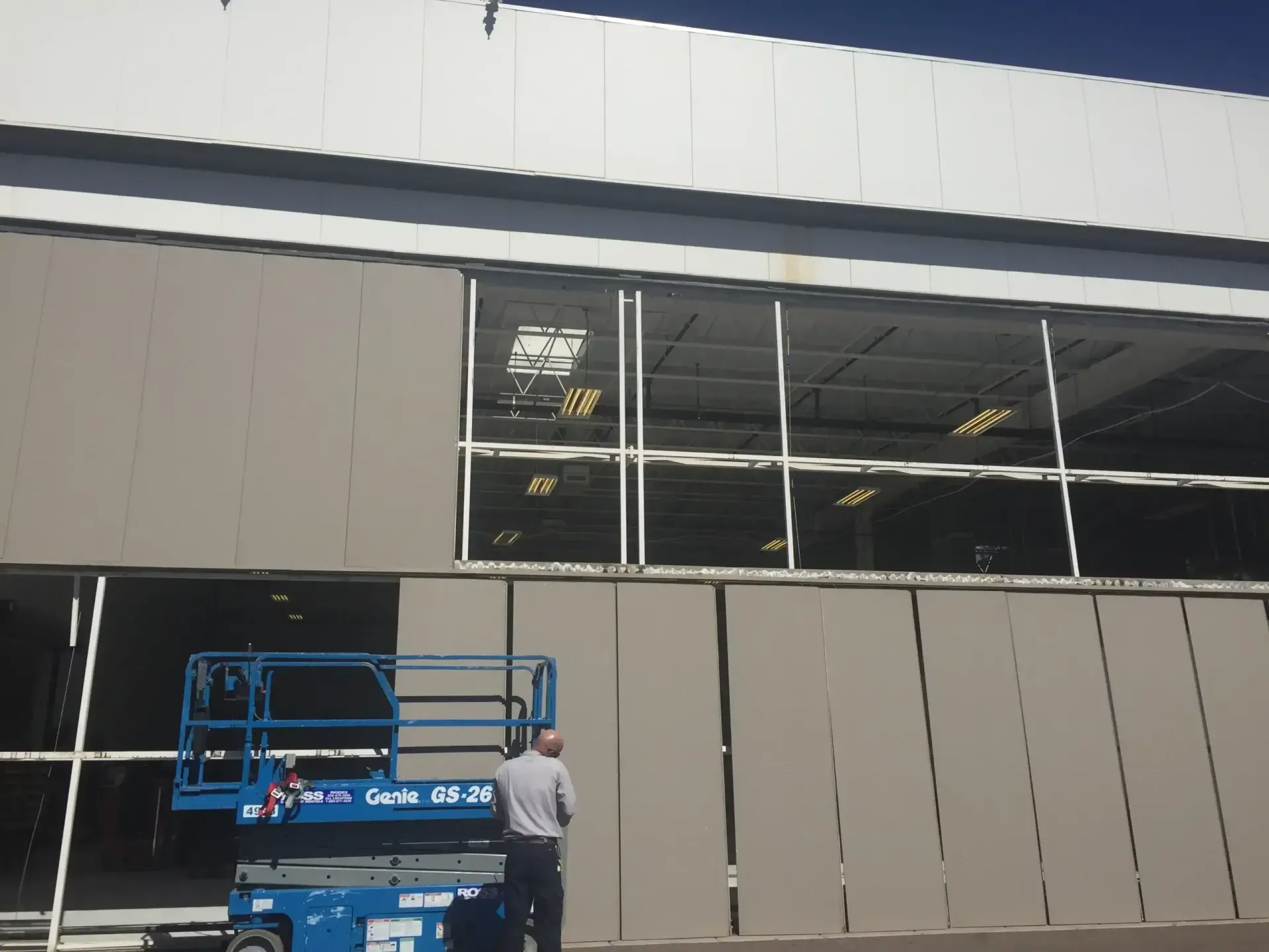 Man on a blue lift working on a light gray industrial building with large windows.