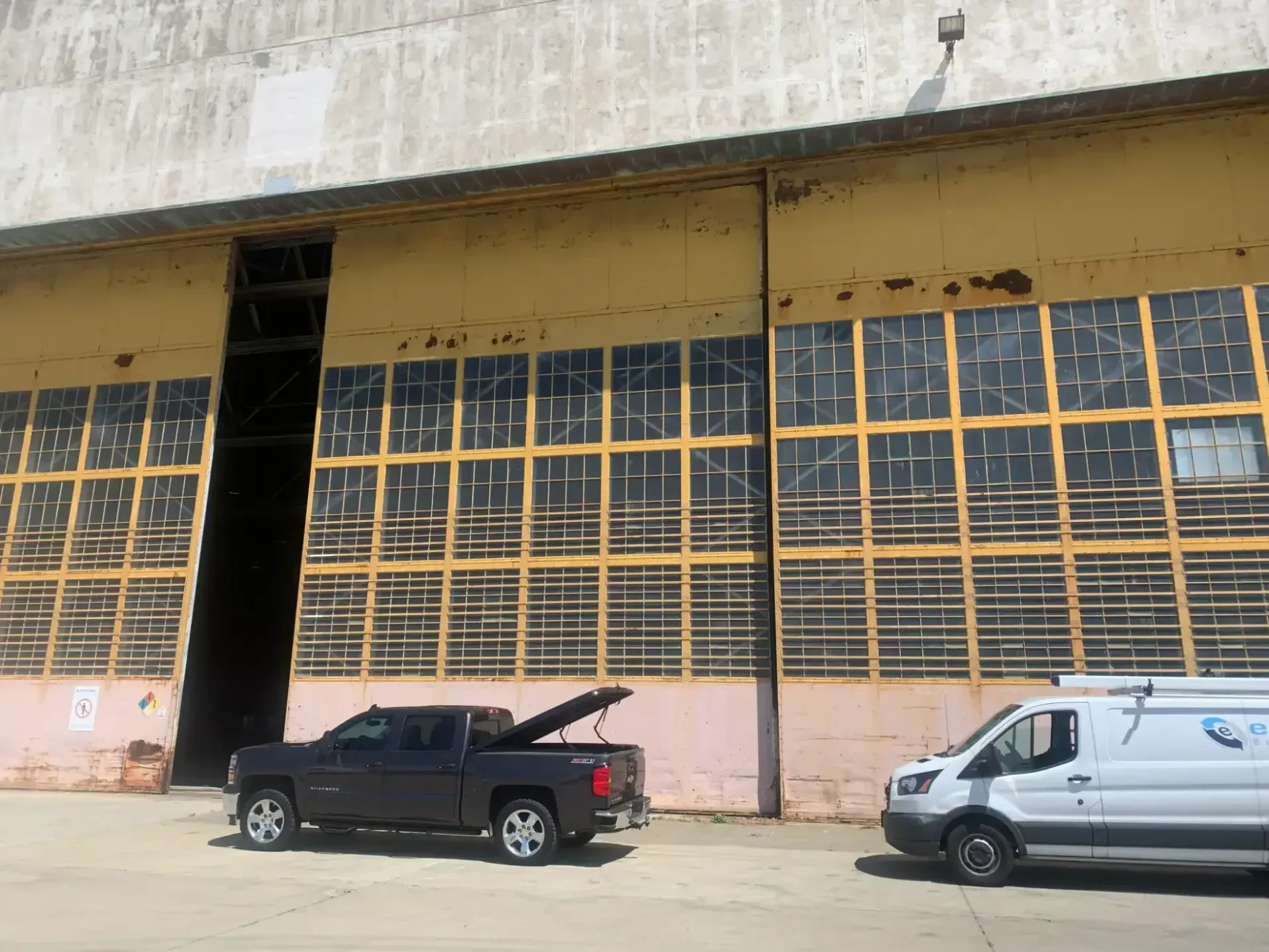 Exterior of a large industrial building with yellow sliding doors; a dark pickup truck and white van are parked in front.