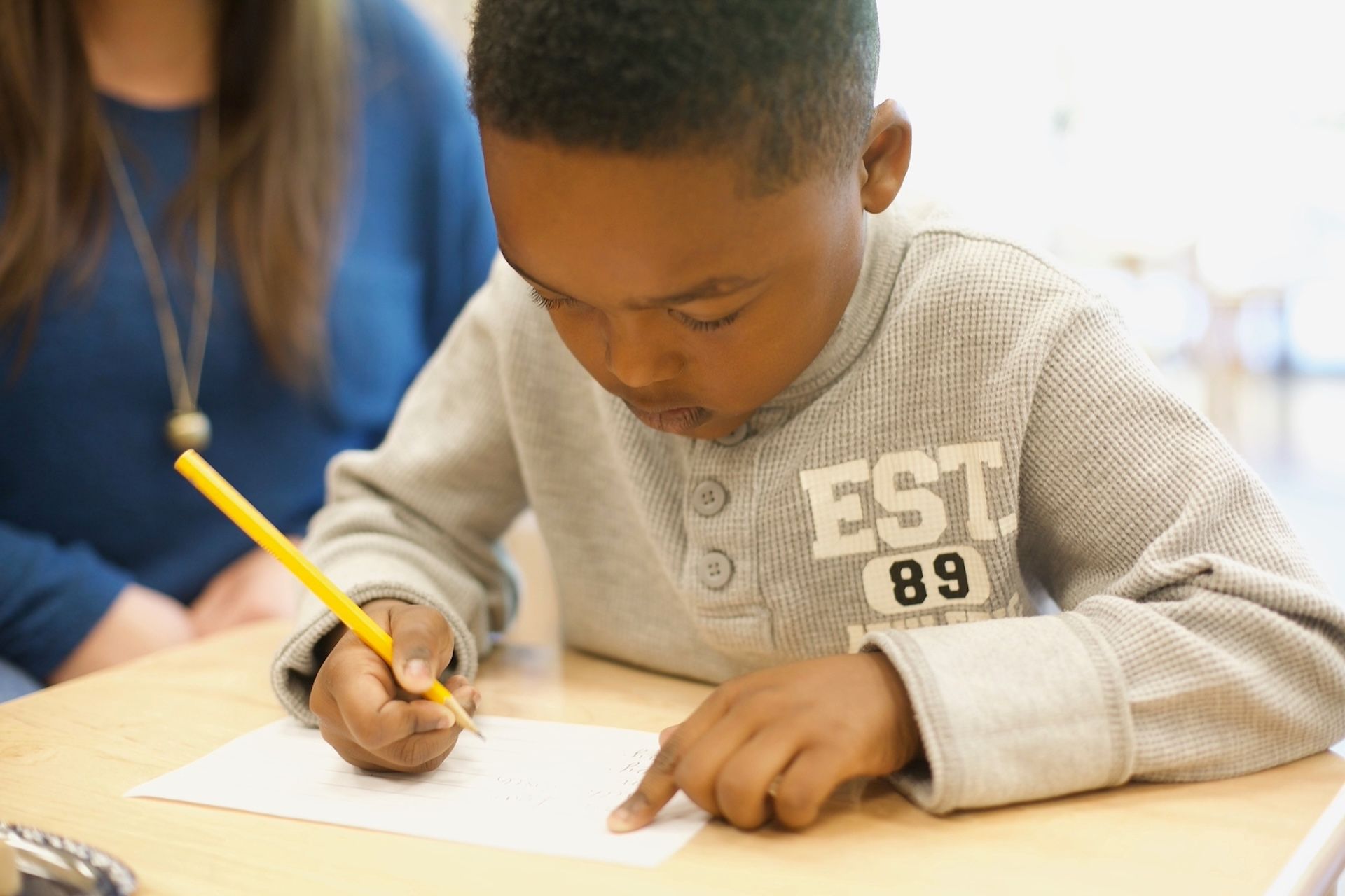 Boy writing at a desk, pointing to paper with pencil in hand. A person sits nearby.