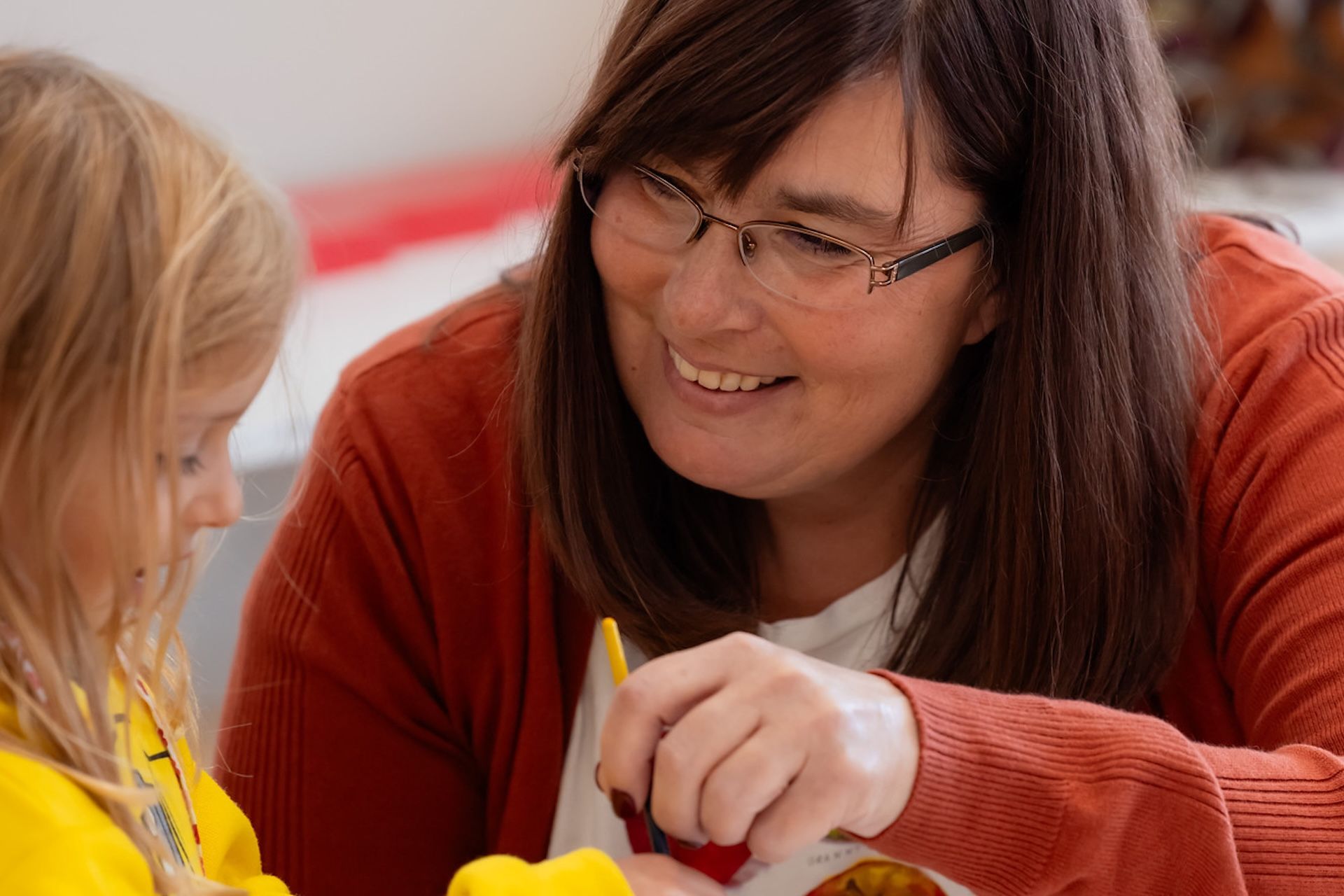 Woman with glasses smiles at a child, both crafting with yellow play dough.