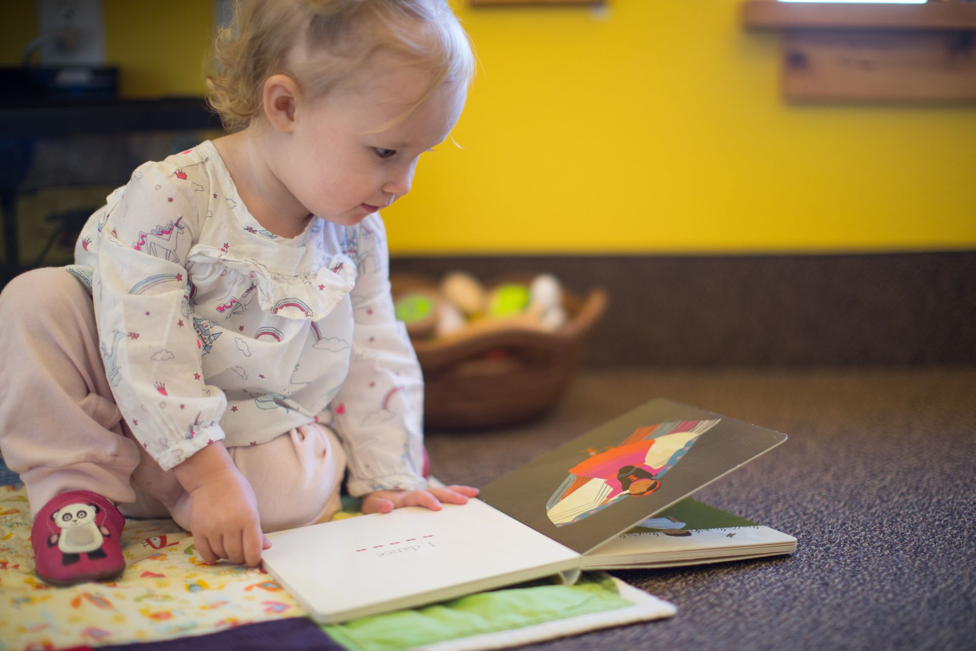 Child sitting on a carpet, looking at an open picture book in a bright classroom.
