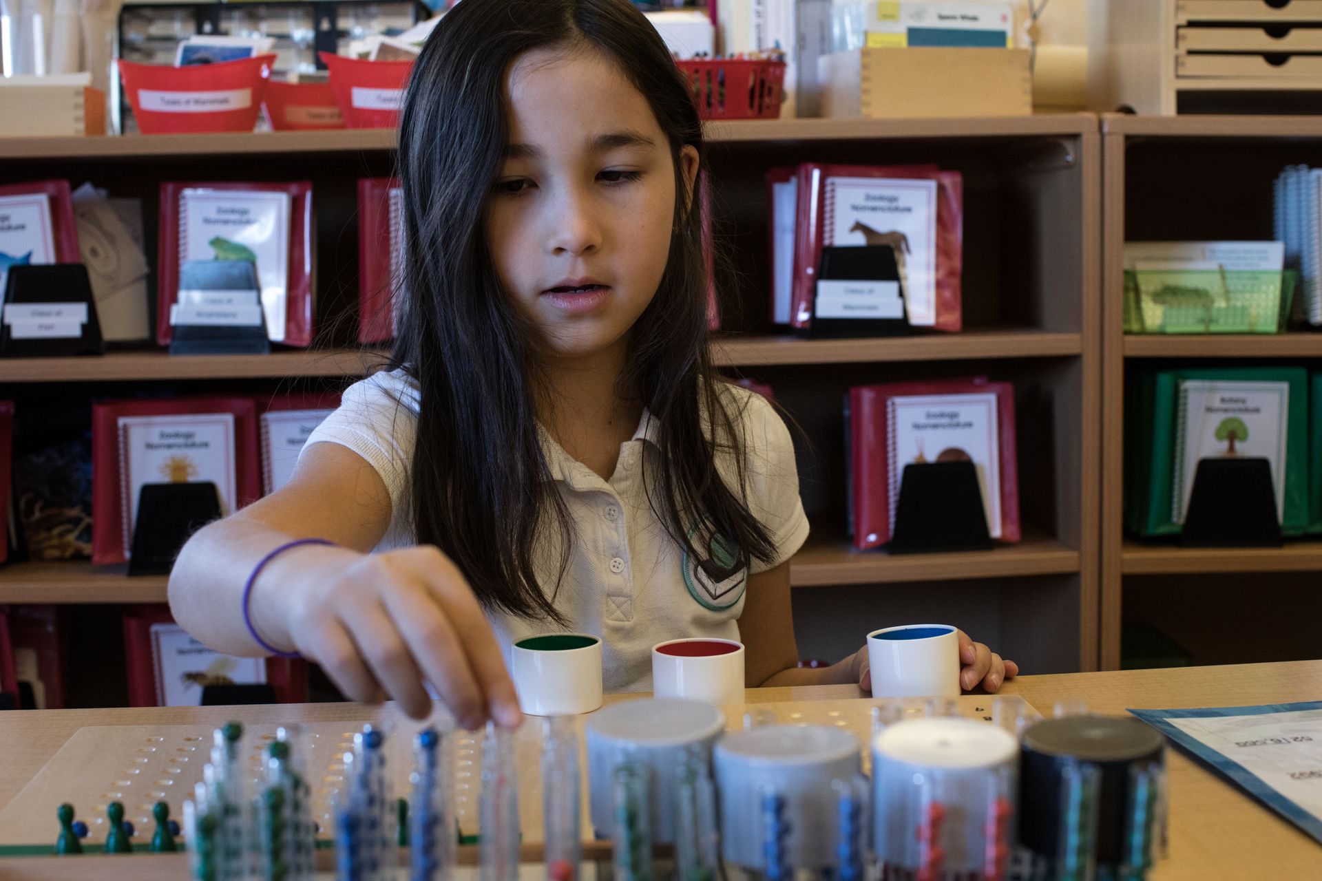 Child arranging colorful bottles on a table in a classroom, with shelves of books behind her
