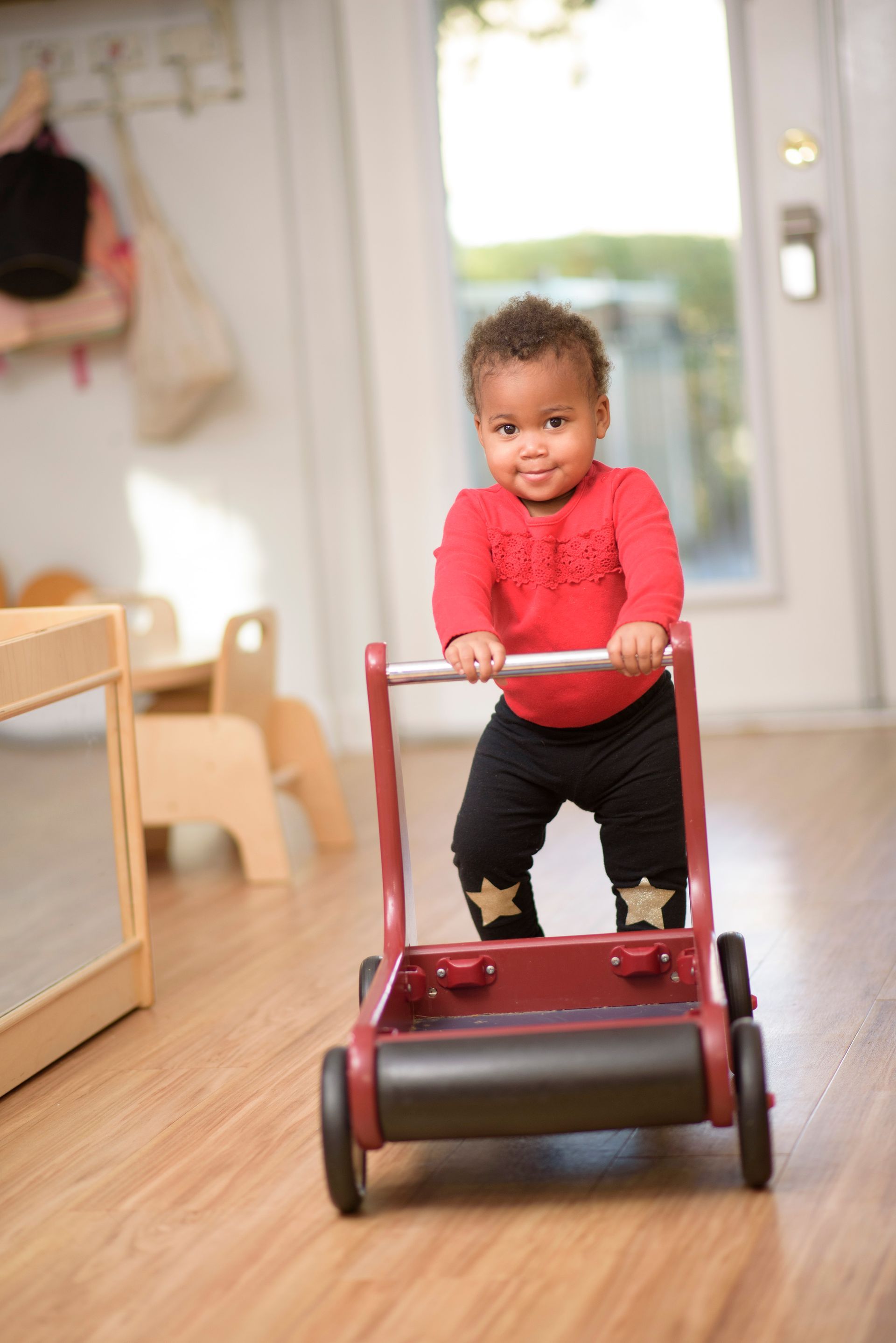 Toddler in a red sweater pushing a red toy walker down a hallway indoors