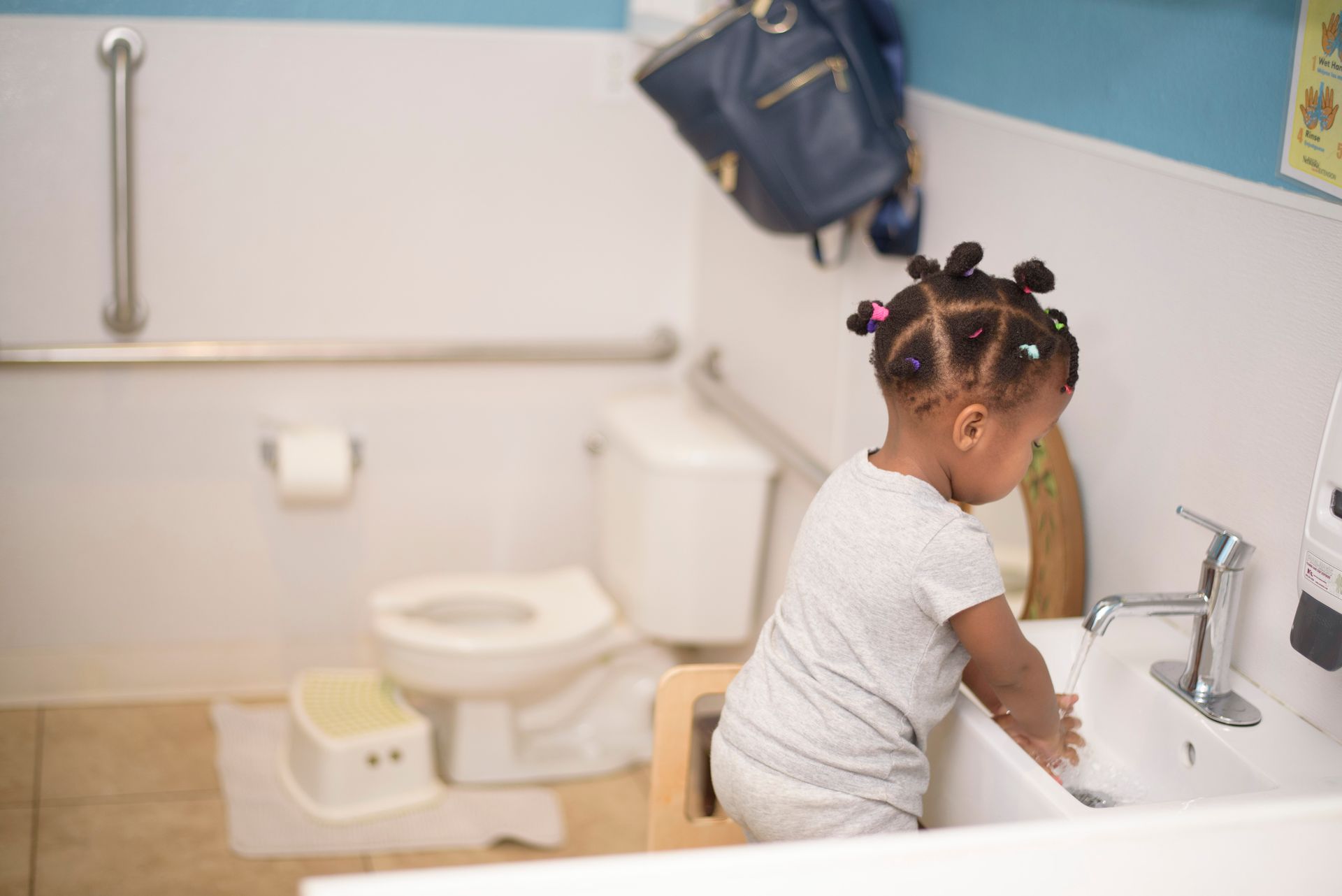 Child washing hands at a bathroom sink beside a toilet and towel hanging on the wall