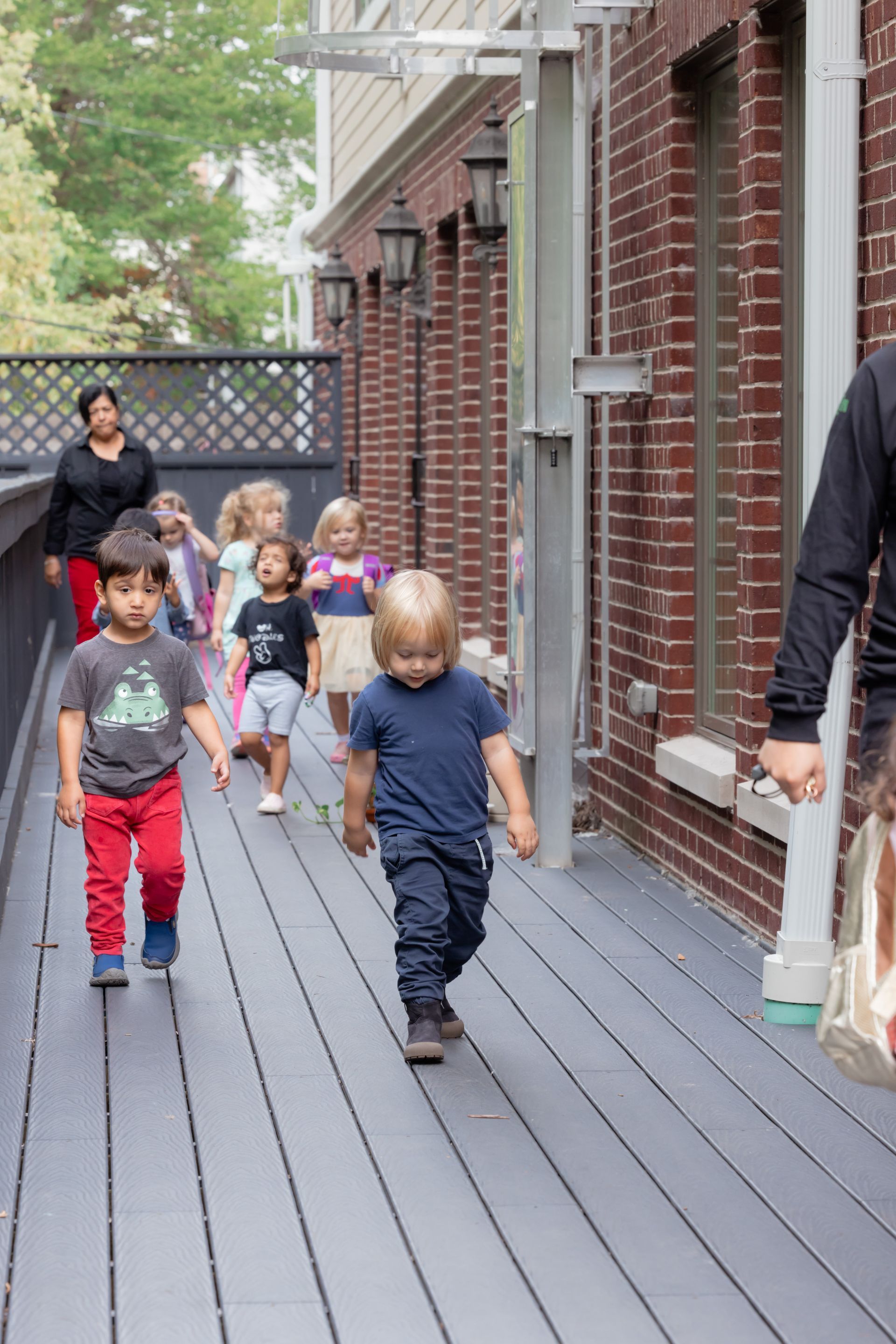 Children walking along a wooden deck beside a brick building, with an adult in the background.