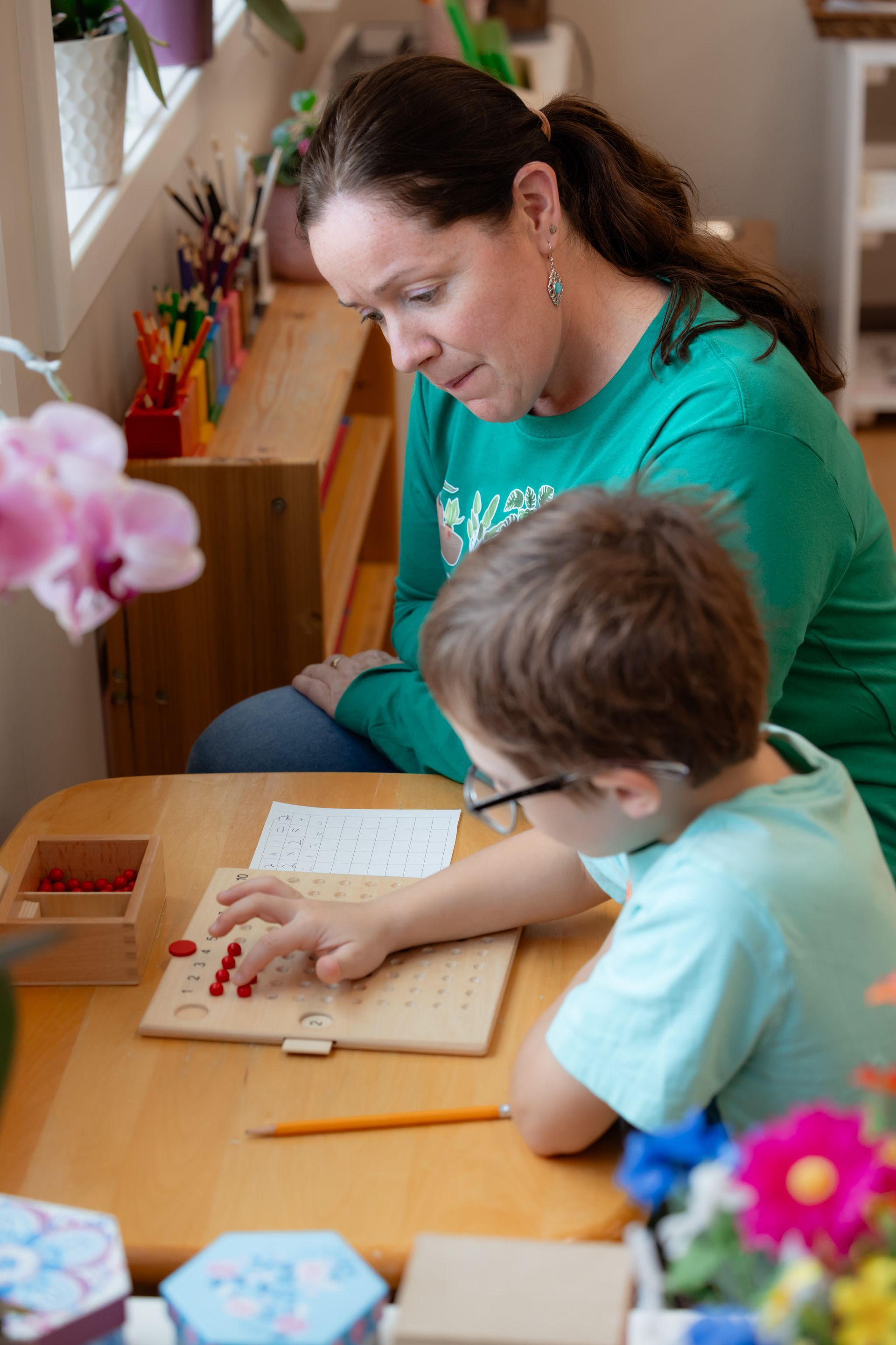 A teacher watches as a young child counts red objects on a wooden table.