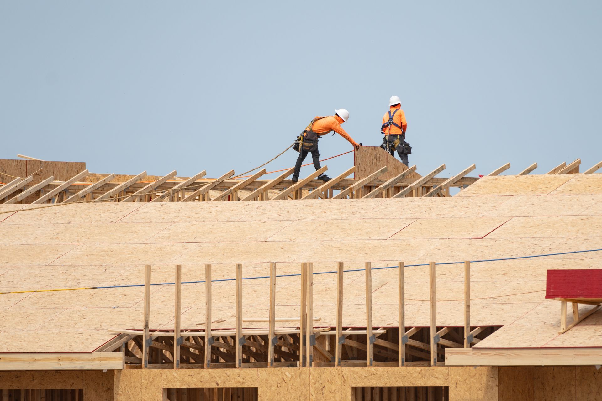 Two construction workers installing plywood on a roof under a blue sky.