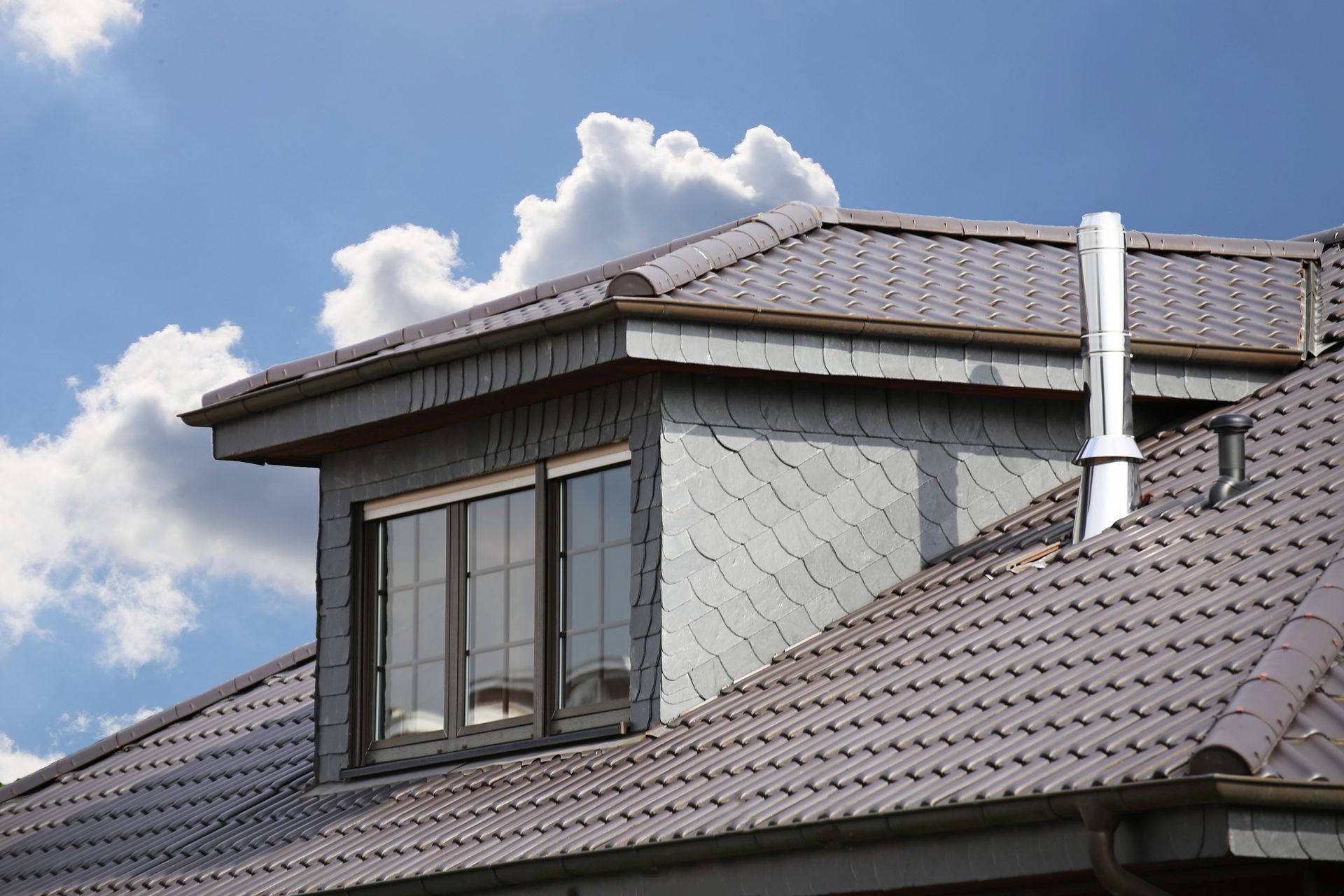Gray tiled roof with a dormer and a chimney against a blue sky with clouds.