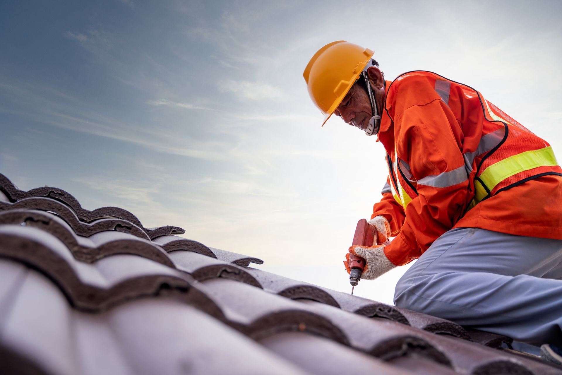 Roofer in orange safety gear using a drill on a tile roof against a cloudy sky.