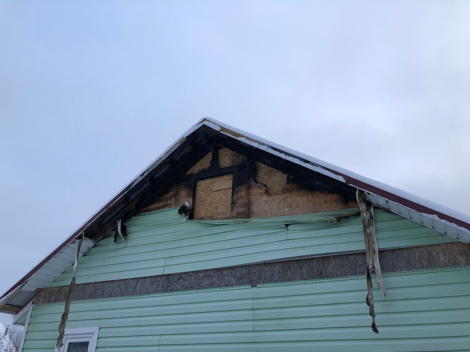 Damaged house gable with charred wood and torn siding against a cloudy sky.
