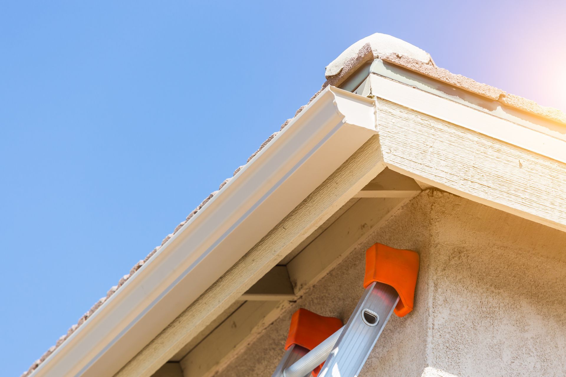Ladder leaning against a house with beige siding, a white gutter, and a clear blue sky.