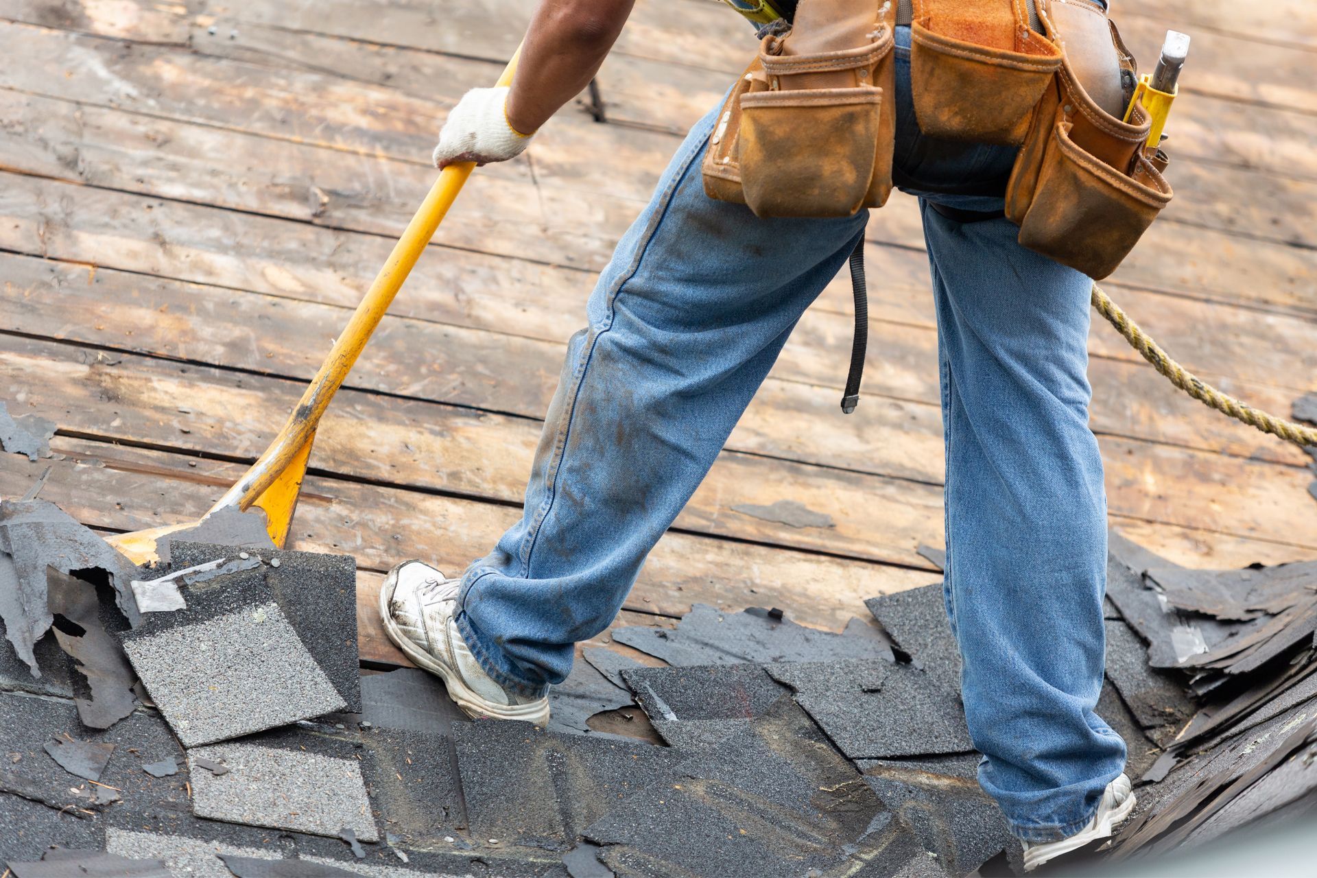 Roofer removing old shingles with a shovel, wearing blue jeans, work boots, and a tool belt.