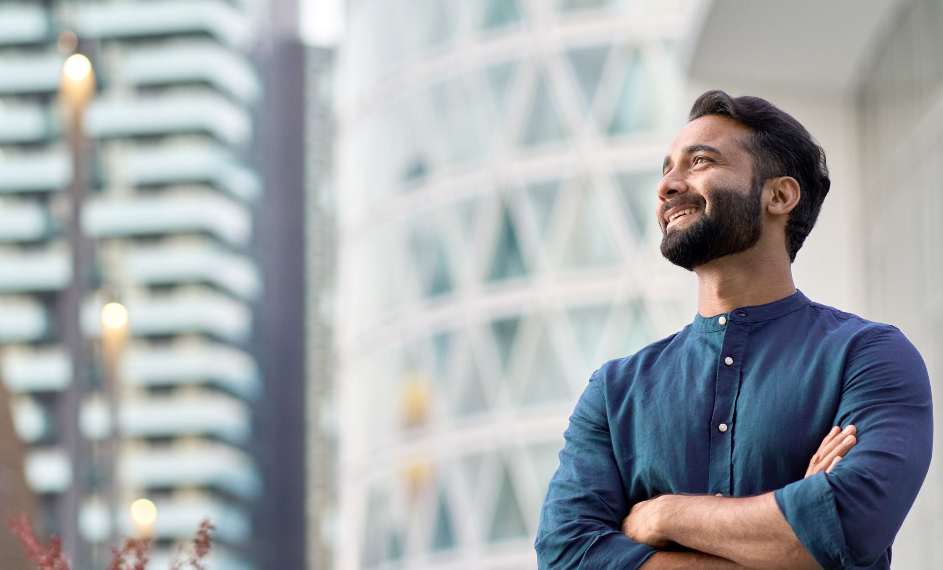 A man with a beard is standing with his arms crossed in front of a building.