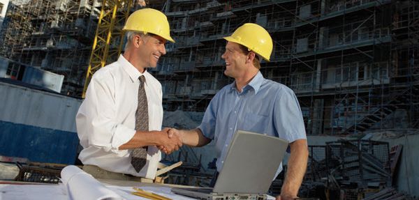 Two men wearing hard hats are shaking hands at a construction site.