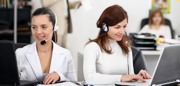 Two women wearing headsets are sitting in front of laptops.