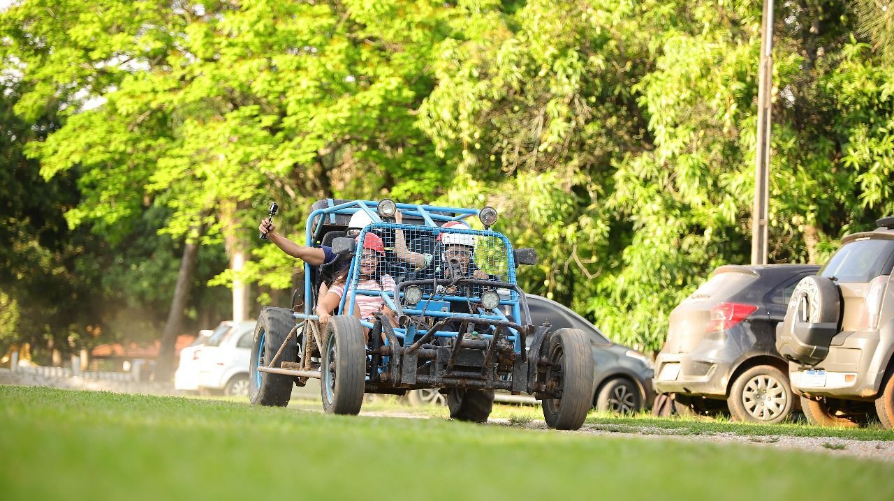 Um carro improvisado, com moldura azul, trafega pela grama, com uma pessoa acenando. Carros estacionados enfileiram-se ao lado.