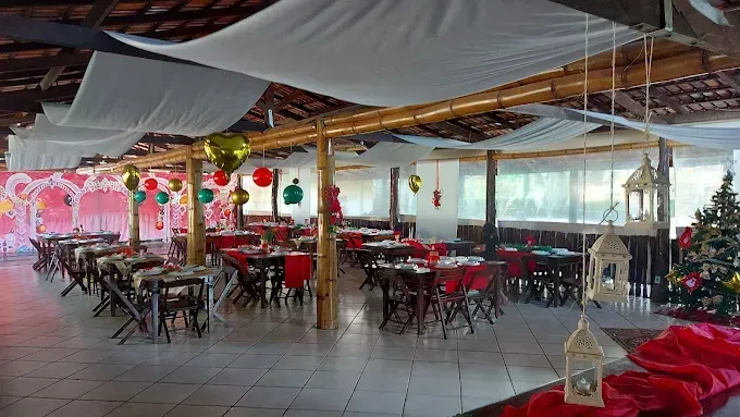 Dentro de um espaço decorado para festas, com mesas postas para refeições. Tecido branco cobre o teto e balões coloridos pendem de vigas de madeira.