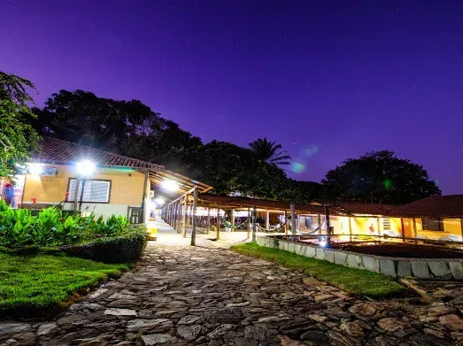 Um caminho de pedras atravessa um complexo de restaurantes ao entardecer. Prédios com interiores iluminados sob um céu roxo.