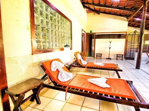 Duas espreguiçadeiras laranjas com toalhas brancas, em um pátio de ladrilhos sob uma área coberta com janelas de vidro.