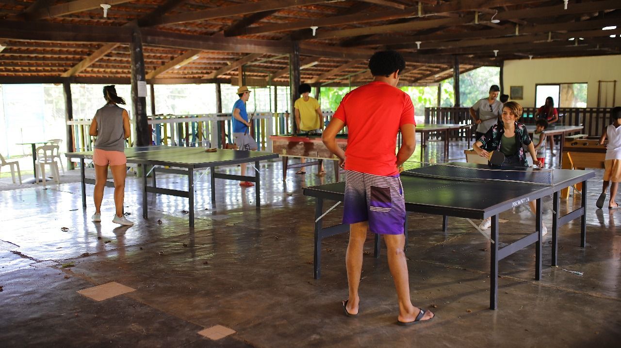 Pessoas jogando tênis de mesa em uma área externa coberta. Várias mesas com jogadores, alguns em trajes casuais, são visíveis.