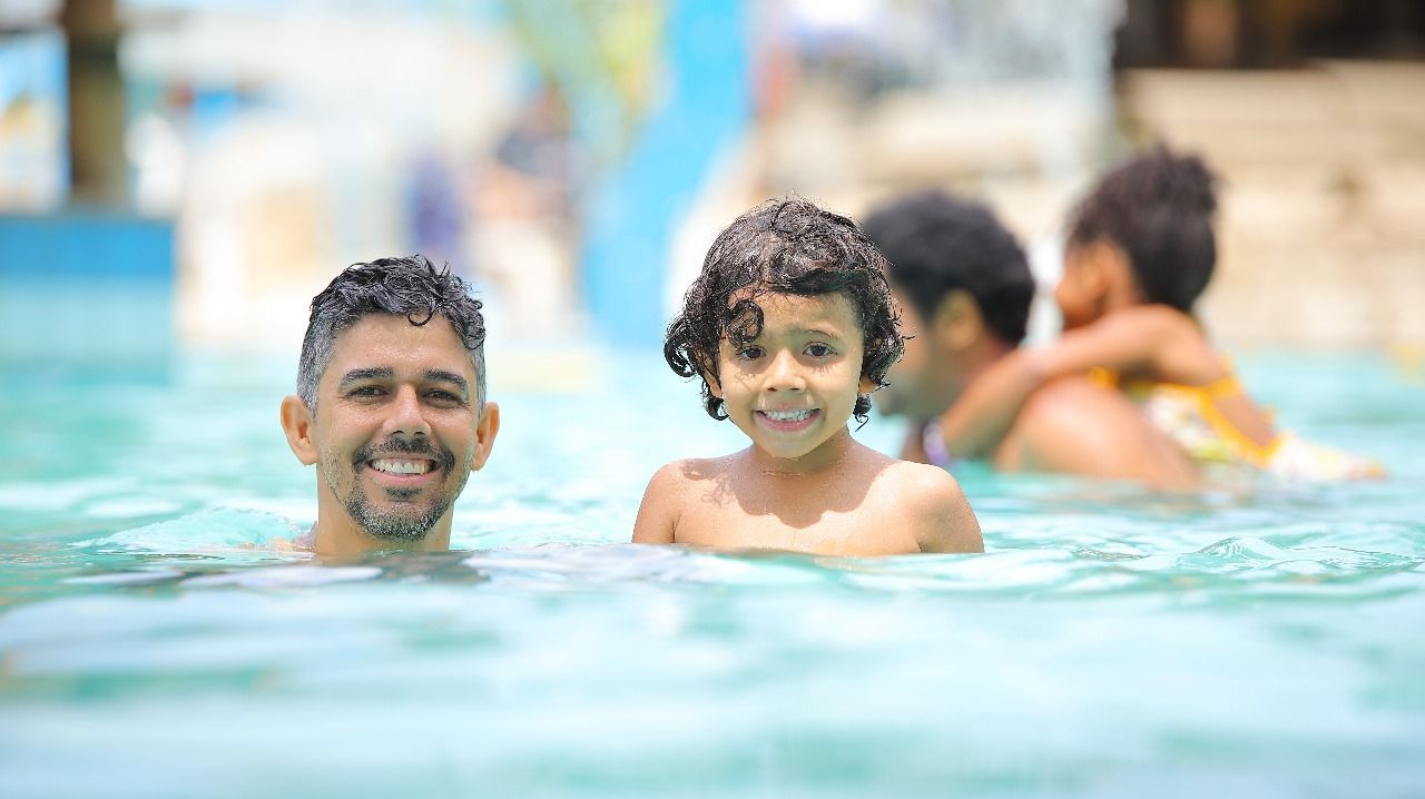Homem e criança sorrindo em uma piscina, outro homem segurando uma criança ao fundo.
