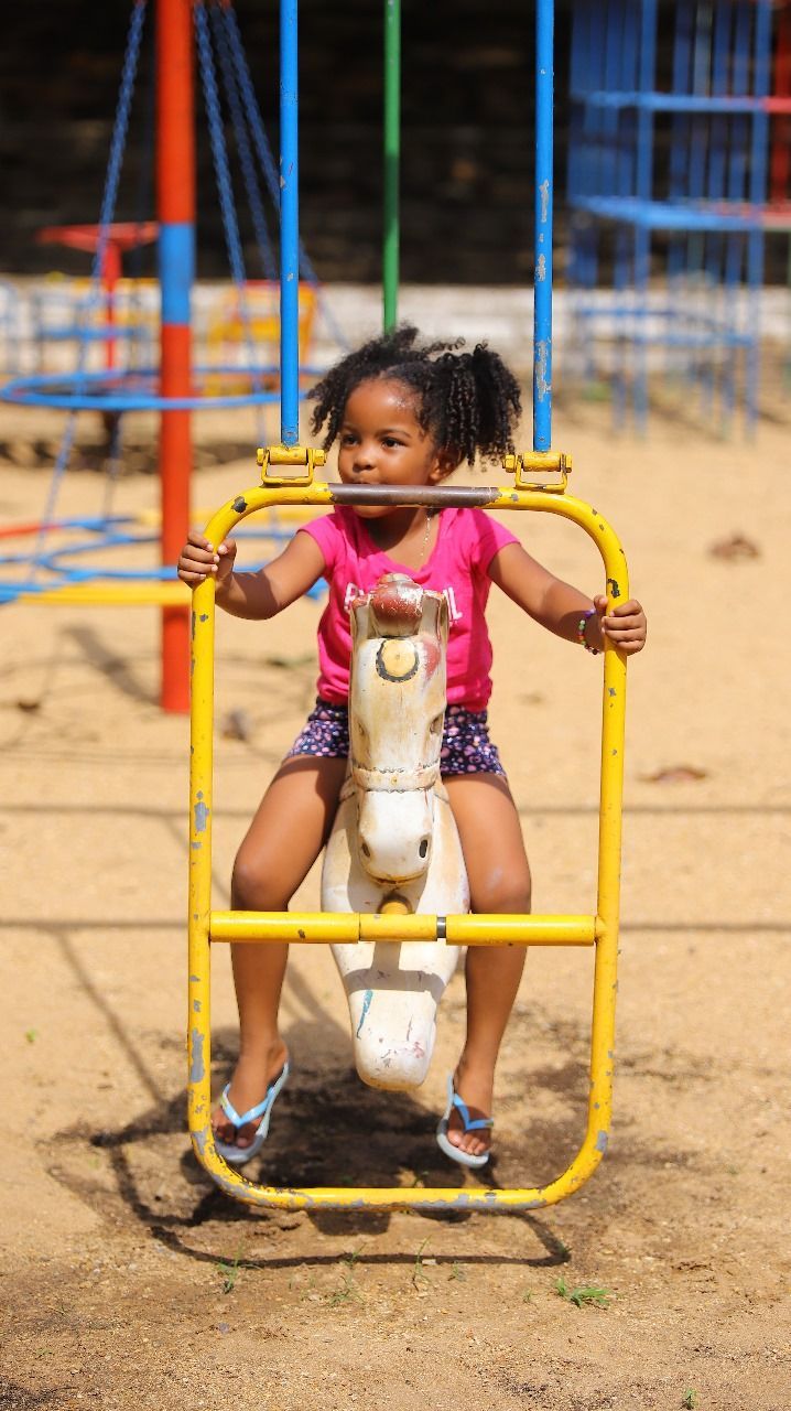 Uma jovem negra monta um cavalo de balanço amarelo e branco em um parquinho. Ela veste uma camisa rosa e shorts escuros, olhando para a direita.