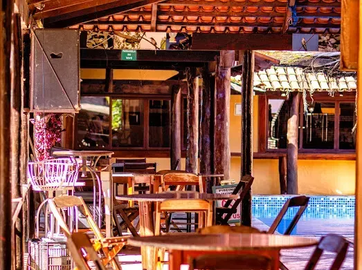Área de jantar ao ar livre com mesas e cadeiras de madeira, com uma piscina visível ao fundo.