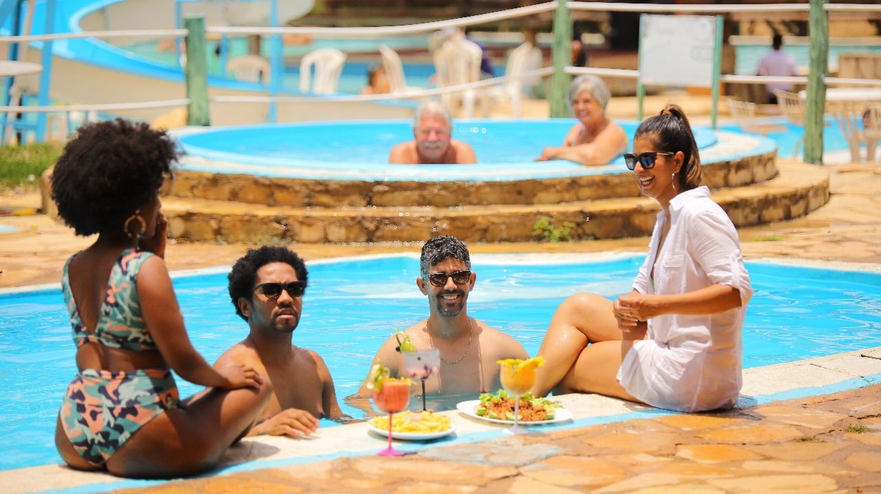 Pessoas relaxando dentro e ao redor de uma piscina com bebidas e petiscos. Duas mulheres e dois homens estão na beira da piscina, enquanto outros nadam nas proximidades.