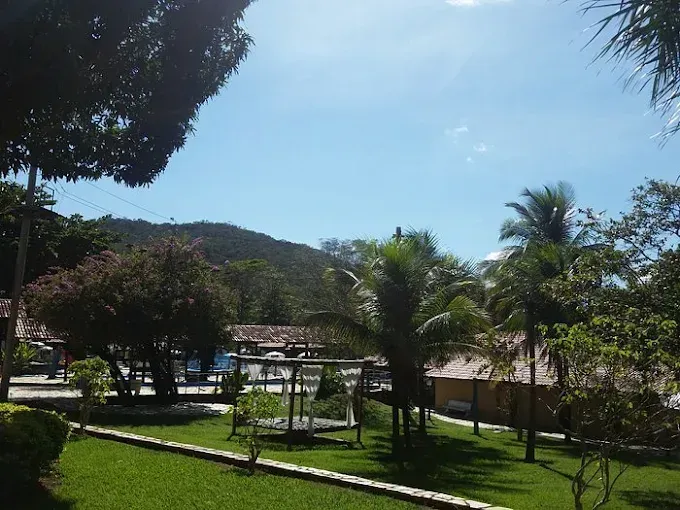 Parque verdejante com árvores, um playground e edifícios sob um céu azul brilhante, com montanhas ao fundo.