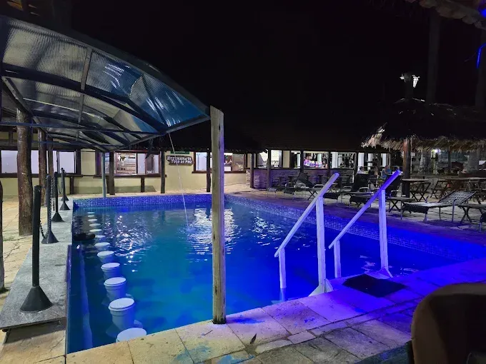 Piscina com iluminação subaquática azul, ao lado de um bar com mesas e cadeiras. Uma passarela coberta é vista acima.
