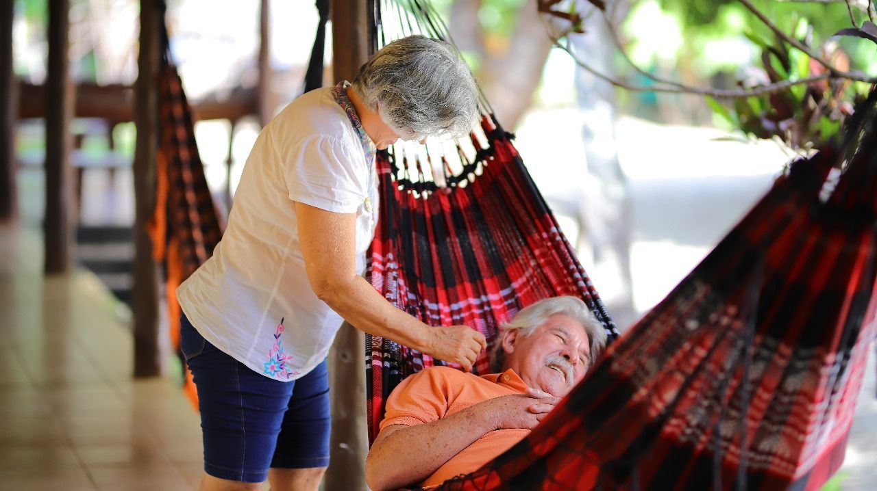Uma senhora idosa ajusta o pescoço de um senhor idoso que relaxa em uma rede vermelha, na varanda.