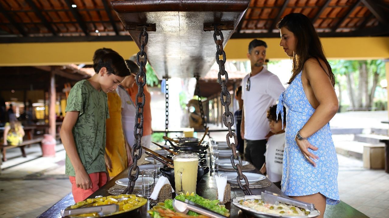 Família em um bufê, observando a comida. O ambiente é ao ar livre, com uma área coberta e mesas de madeira.