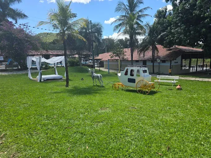 Gramado verdejante com um canteiro branco e uma carruagem amarela. Palmeiras e prédios ao fundo, sob um céu azul.