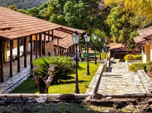 Fileira de prédios com telhados e passarelas marrons. Um caminho de pedras ladeado por grama verde e postes de luz. Uma vegetação exuberante circunda o local.