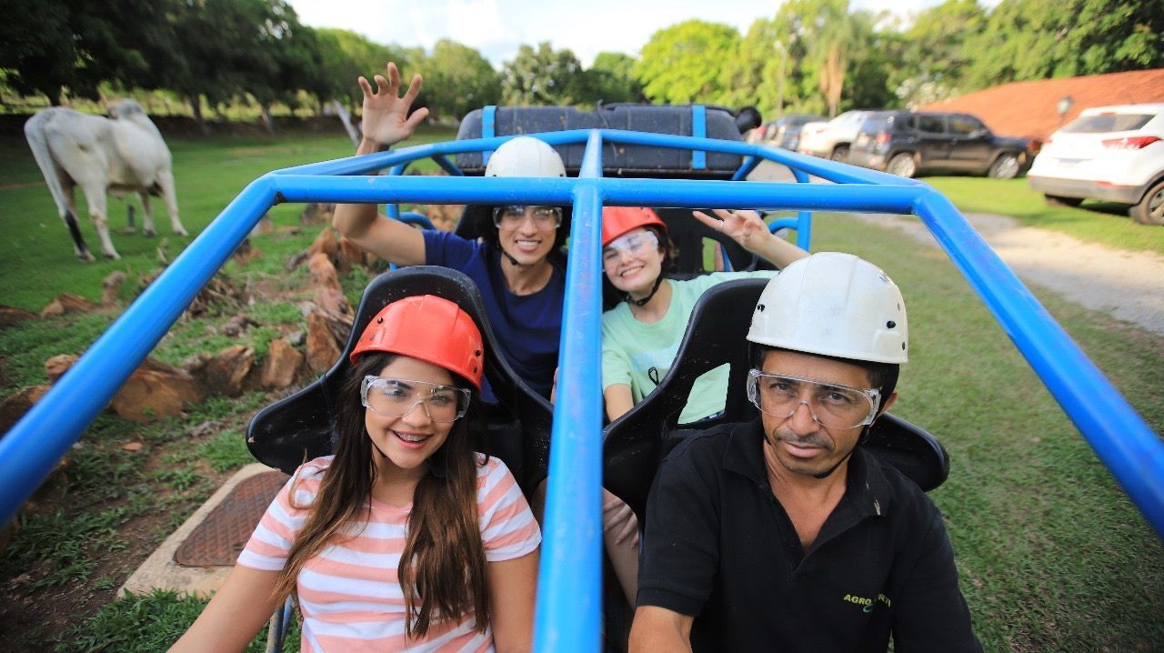 Pessoas em um carrinho azul passeiam por uma área gramada, usando capacetes e óculos de segurança, acenando para a câmera. Uma vaca pasta nas proximidades.