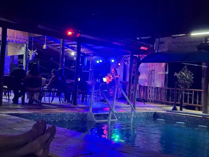 Pés em primeiro plano com vista para uma piscina com luzes azuis, levando a uma área externa coberta com pessoas e iluminação adicional à noite.