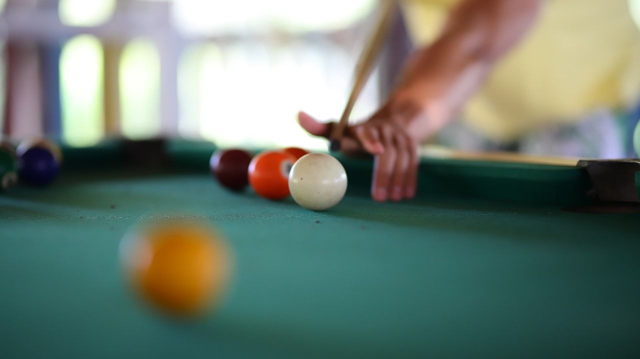 Pessoa alinhando uma tacada de sinuca em uma mesa de feltro verde. A bola branca está em foco, com outras bolas coloridas ao fundo.