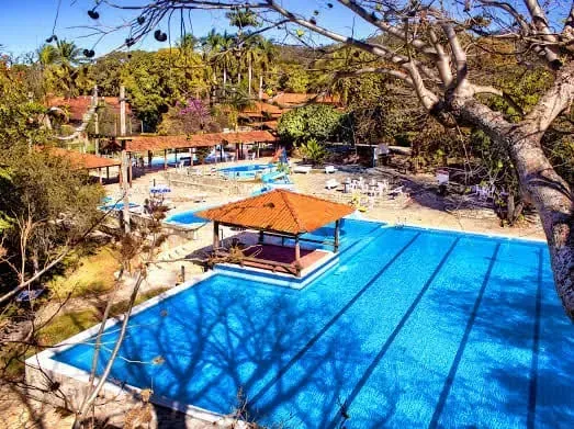 Vista de cima de uma área ensolarada de piscina externa com um gazebo e várias piscinas. Árvores e prédios ao fundo.