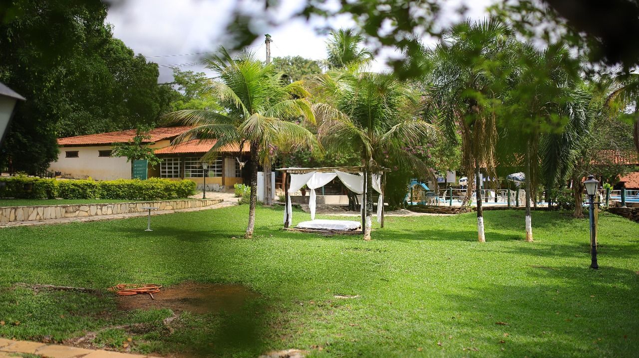 Gramado verdejante com um gazebo branco, cercado por árvores. Ao fundo, vê-se um prédio com telhado marrom.