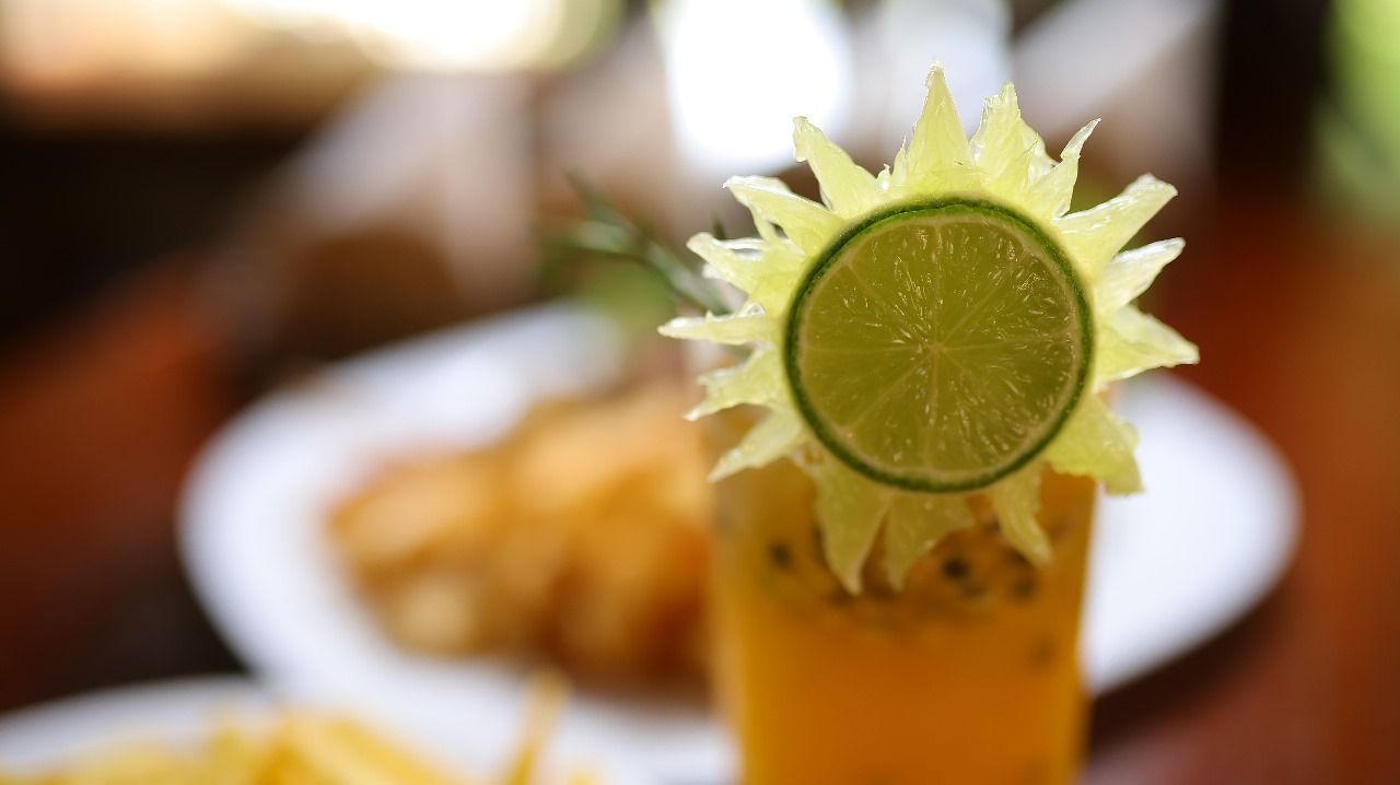 Coquetel decorado com uma fatia de limão cortada para representar o sol. Coquetel amarelo em um copo alto com um prato de comida desfocado ao fundo.