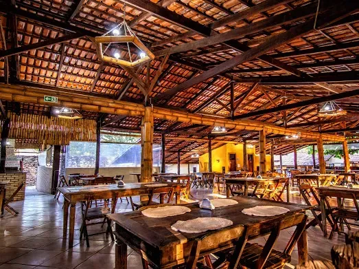 Interior rústico de restaurante com mesas de madeira, cadeiras e telhado de telhas. A luz natural entra pelas laterais abertas.