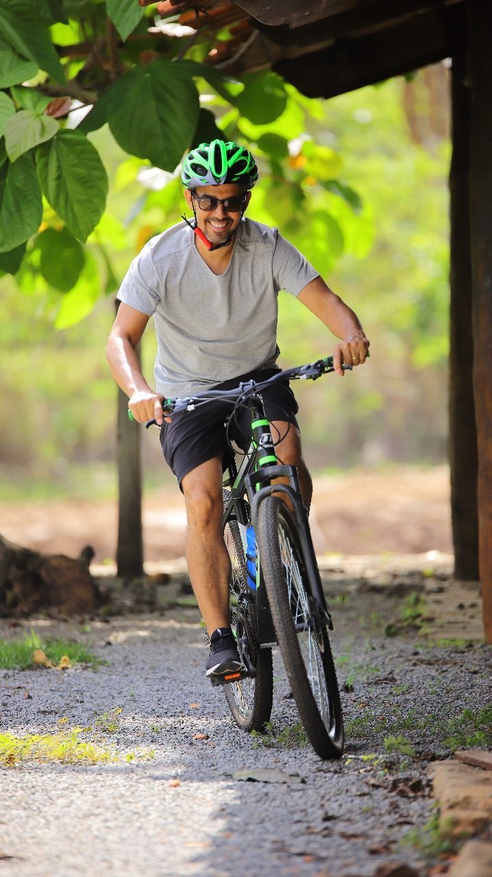 Homem de camisa cinza e capacete verde sorri enquanto anda de mountain bike em um caminho de cascalho sombreado por árvores.